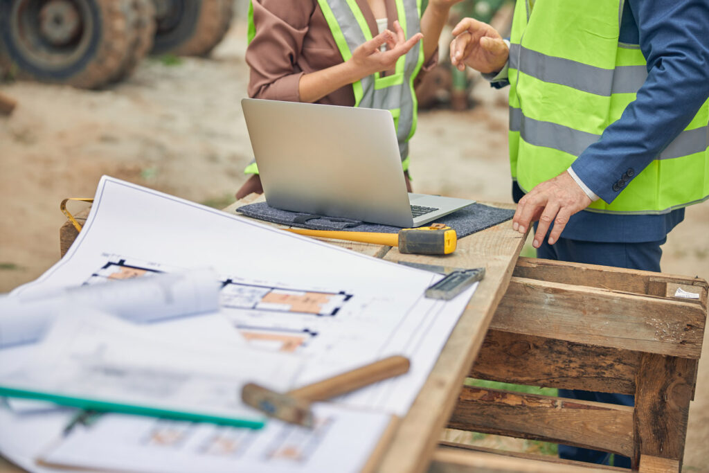 Ingénieurs en discussion sur chantier avec plans et ordinateur portable.